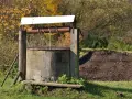 Stara studnia kręgowa z drewnianym kołowrotem i metalową korbą, otoczona jesienną zielenią i stertą ziemi.