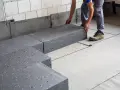 Pracownik układa szary styropian pod podłogówkę. Elementy tworzą izolację na betonowym podłożu.