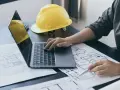 Architekt analizuje plany, porównując powierzchnię użytkową z całkowitą. Na biurku leży żółty kask i laptop.