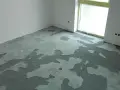 Nierównomiernie rozłożona, mokra krzywa wylewka samopoziomująca na podłodze w rogu pomieszczenia, obok okna.