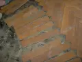 Drewniana podłoga w jodełkę, częściowo rozebrana. Widać luźne deseczki i pył. Subit co to?