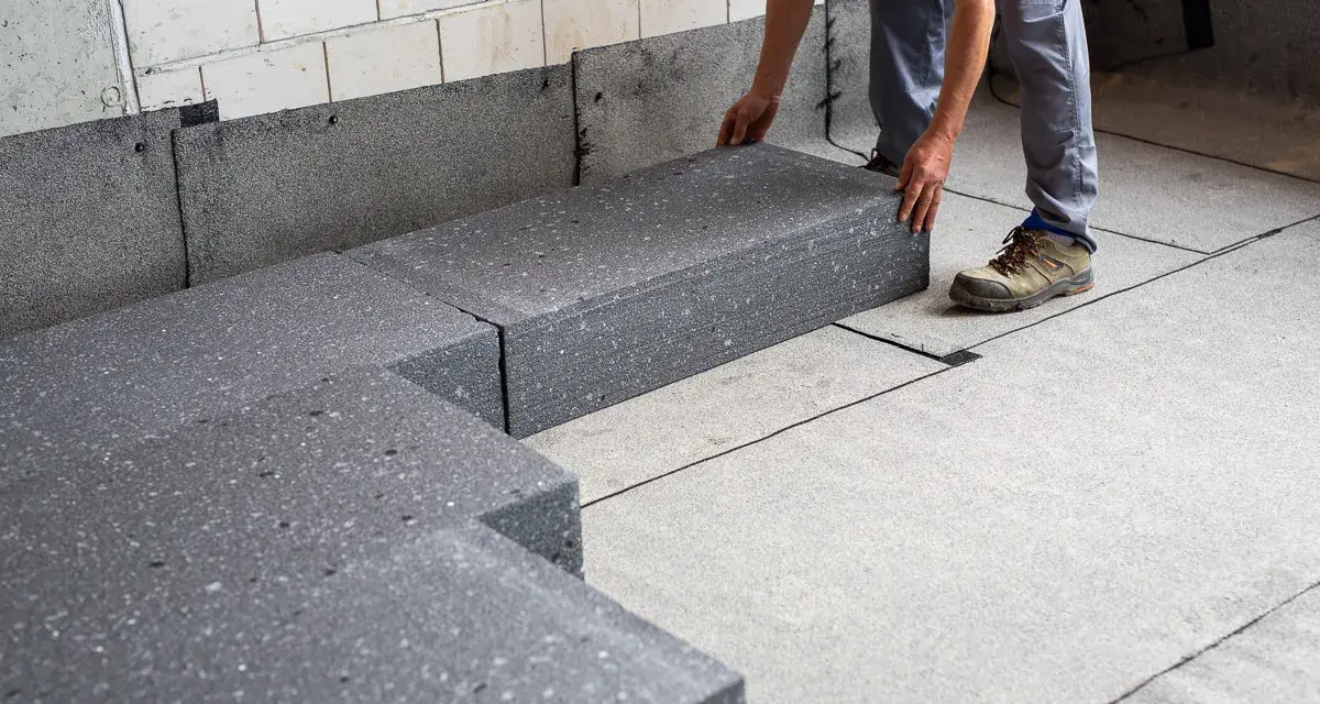 Pracownik układa szary styropian pod podłogówkę. Elementy tworzą izolację na betonowym podłożu.