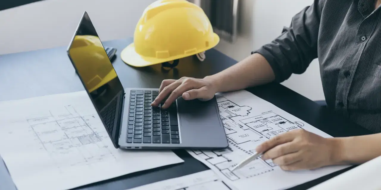 Architekt analizuje plany, porównując powierzchnię użytkową z całkowitą. Na biurku leży żółty kask i laptop.