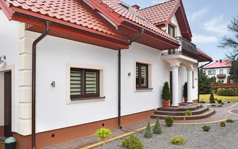 Elegancki dom z białym tynkiem i brązowymi detalami. Cokół wykończony ozdobnym tynkiem mozaikowym dodaje mu charakteru.