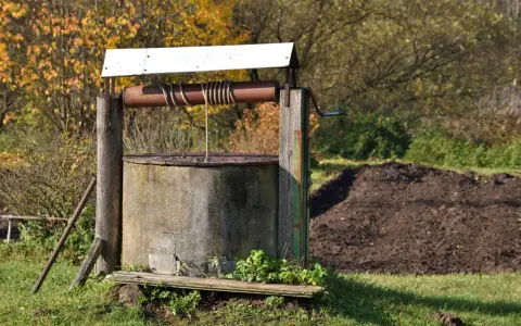 Stara studnia kręgowa z drewnianym kołowrotem i metalową korbą, otoczona jesienną zielenią i stertą ziemi.