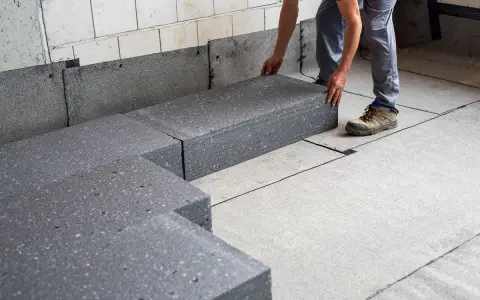 Pracownik układa szary styropian pod podłogówkę. Elementy tworzą izolację na betonowym podłożu.
