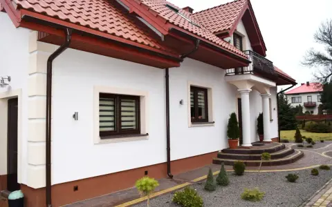 Biały dom z brązowym cokołem wokół, czerwonym dachem i ozdobnymi schodami.