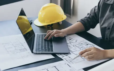 Architekt analizuje plany, porównując powierzchnię użytkową z całkowitą. Na biurku leży żółty kask i laptop.