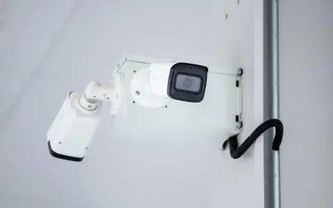 Dwie kamery monitoringu na białej ścianie. Zastanawiasz się, gdzie zgłosić nielegalny monitoring?