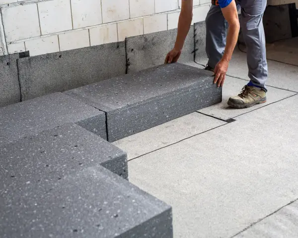 Pracownik układa szary styropian pod podłogówkę. Elementy tworzą izolację na betonowym podłożu.