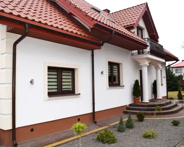 Biały dom z brązowym cokołem wokół, czerwonym dachem i ozdobnymi schodami.