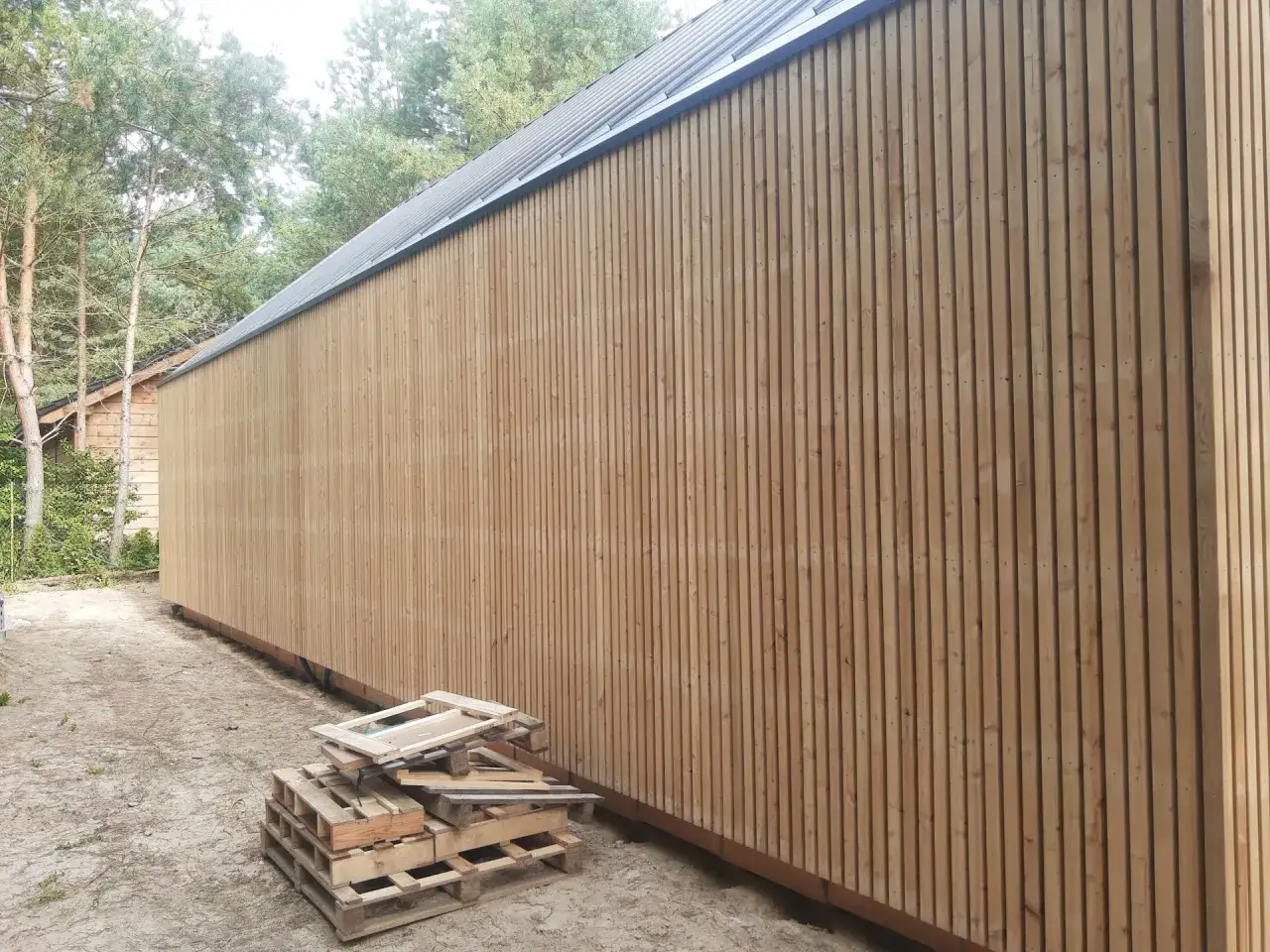 Nowe deski elewacyjne tworzą nowoczesne aranzacje. Drewniana ściana budynku z metalowym dachem, obok palety.