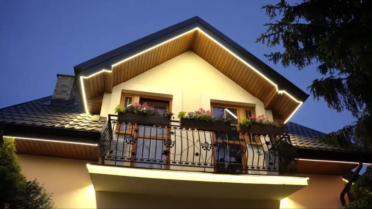 Elewacja domu z podbitką drewnianą, ozdobiona ciepłym oświetleniem LED. Balkon z kutą balustradą i kwitnącymi skrzynkami.