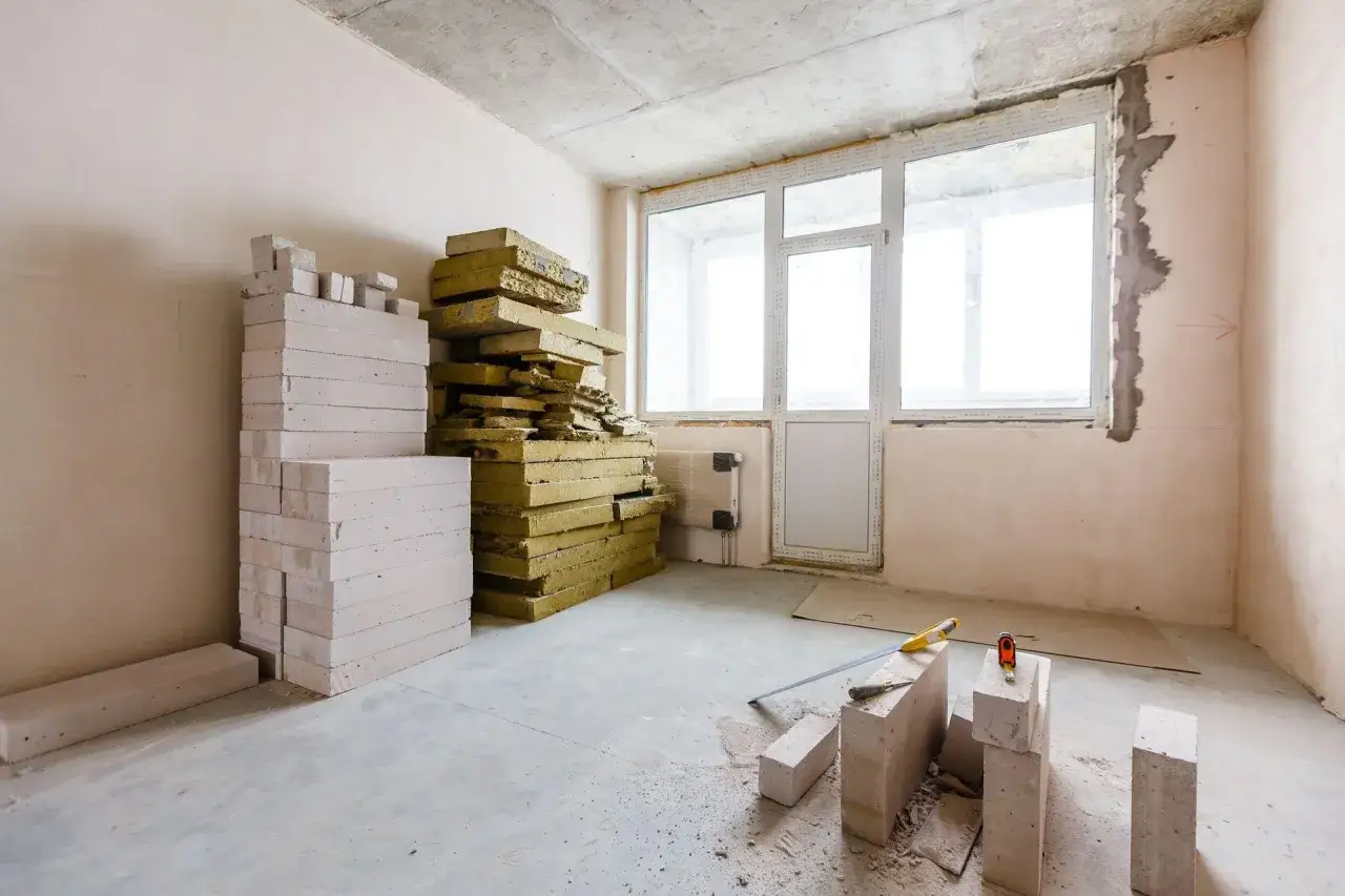 Materiały budowlane, w tym pustaki i wełna mineralna, czekają na ocieplanie od wewnątrz. W pomieszczeniu widać okno i drzwi.