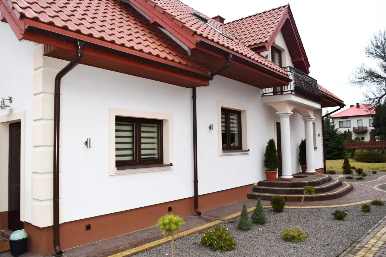 Nowy dom z białym tynkiem i brązowym dachem. Wokół domu widać zadbany ogród z kamyczkami i roślinnością. Elegancki cokół wokół domu dodaje mu uroku.