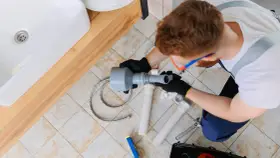 Hydraulik pracuje nad odpowietrzeniem pionu sanitarnego, zgodnie z normą. Widoczny jest zlew i narzędzia.