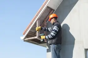 Budowlaniec montuje podbitkę ze styropianu, przykręcając drewniane elementy do okapu domu.