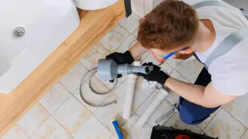 Hydraulik pracuje nad odpowietrzeniem pionu sanitarnego, zgodnie z normą. Widoczny jest zlew i narzędzia.