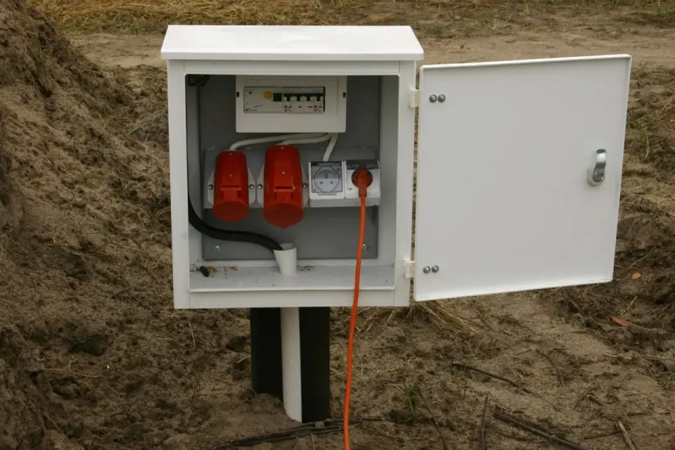 Otwarta skrzynka elektryczna na działce z bezpiecznikami i gniazdami. Pomarańczowy kabel wychodzi z gniazda, sugerując dalsze podłączenia.
